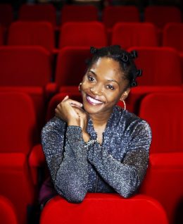 Profile photo of Tamar Dixon, a young black woman seated in a theatre surrounded by red velvet seats