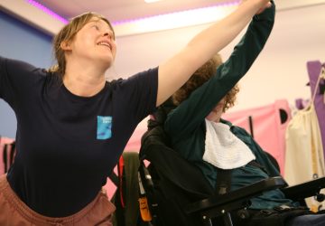 A dance artist and a young lady in a wheel chair hold arms outstretched whilst dancing