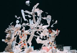 A group of dancers dressed in white against a black backdrop, throwing pieces of white paper in the air