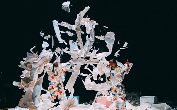 A group of dancers dressed in white against a black backdrop, throwing pieces of white paper in the air