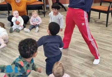 An intergenerational dance session, a lady dances with arms wide in front of a group of small children, older adults looking on
