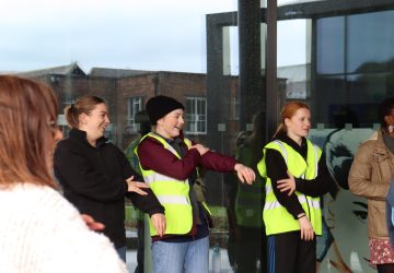 Two young people (Young Producers) looking on at an event, dressed in high vis jackets