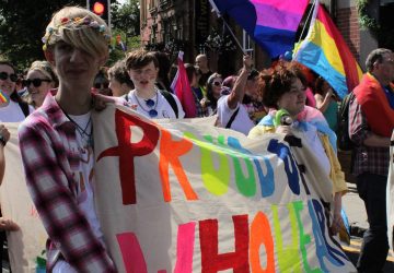 A group of young people at Chester Pride parade with a colourful banner stating "Proud of who we are"