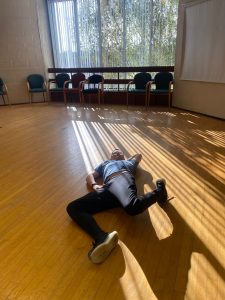 A young adult lying on the floor of a dance studio, legs in a running position. Sun shines through the window creating a lined pattern where it reflects from the blinds