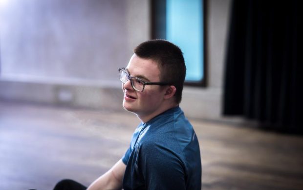 A young adult with Downs Syndrome and glasses sitting on a studio floor. Only the upper body is visible with half his face angled forwards. A slight smile on his face.