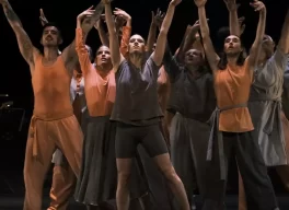 A group of dancers on a darkened stage with their arms in the air. Dancers are dressed in either orange or brown.