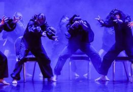A blue-tinged photo of a group of dancers in a row in front of chairs, leaning over with head tucked in and arms outstretched.