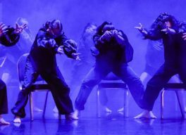 A blue-tinged photo of a group of dancers in a row in front of chairs, leaning over with head tucked in and arms outstretched.